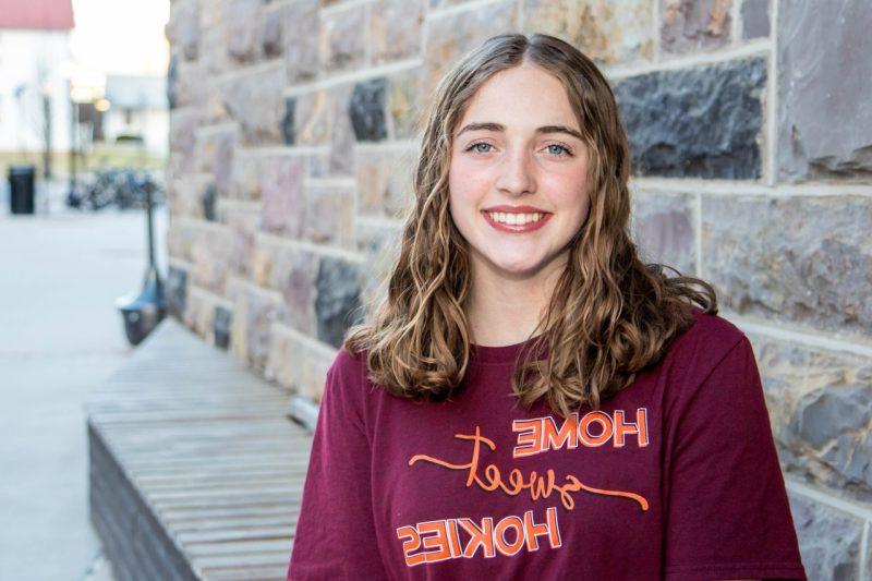 Kirsten Perrin wears a maroon shirt that reads "Home Sweet Hokies" as she sits for a portrait in front of a Hokie Stone wall. 